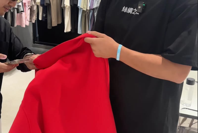 Checking a finished red hoodie by hand while the supplier pulls up its details on a phone